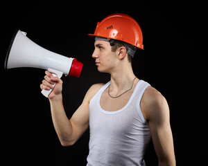 A teenager in an orange construction helmet with a loudspeaker in his hand on a dark background shouts into a megaphone