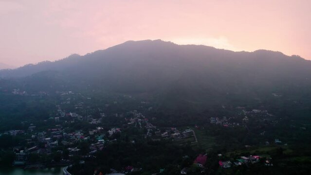 aerial drone shot with sunrise behind huge mountain and small colorful town at the base hidden by the fog showing the natural beauty of uttarakhand cities like nainital, bhimtal, almora and more in