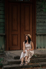 Fototapeta premium woman in beige dress and straw hat is sitting on the stairs and smiling behind the wooden door