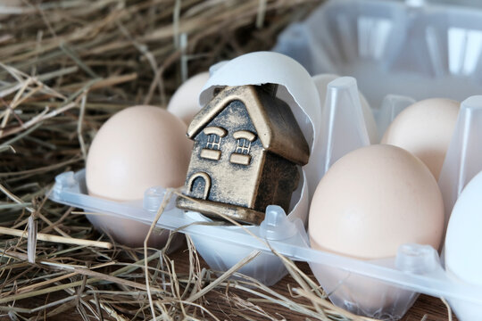 The house is a gilded toy, in an eggshell, white eggs next to a background of straw, hay. The concept of the acquisition of housing, construction, mortgage in a crisis. Easter decoration.