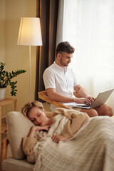Vertical selective focus shot of young adult Caucasian man sitting in living room using laptop while his girlfriend taking nap