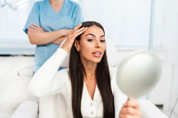 Beautiful and happy young woman sitting in medical chair and looking in the mirror. She is...
