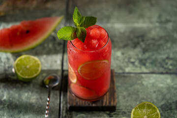 Watermelon lime refreshing cocktail in modern glass (mojito, lemonade, mocktail) with fresh mint. Cold summer fruit drink with beautiful dark grey textured background. Selective focus, copy space.