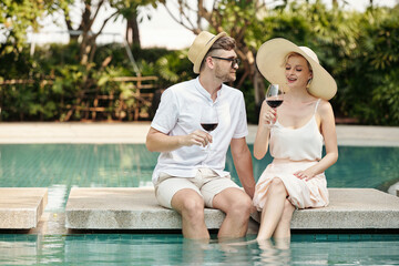 Fototapeta na wymiar Stylish young Caucasian man and woman in love sitting relaxed at pool with legs in water flirting and drinking red wine during vacation