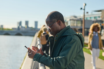 African mature man in eyeglasses typing a message on his mobile phone standing outdoors in the city