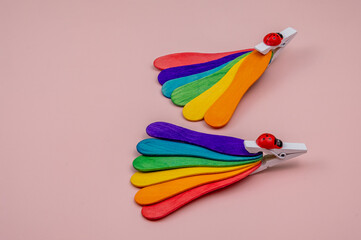Two rainbow fans attached with a white clip with a ladybug on a pink table. Pride day. 
