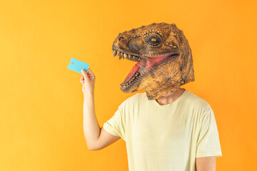 Portrait of young woman in dinosaur animal head mask holding credit card on orange background