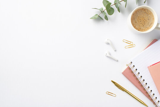 Business concept. Top view photo of workplace stack of pink planners gold pen clips eucalyptus cup of coffee and wireless earbuds on isolated white background with copyspace