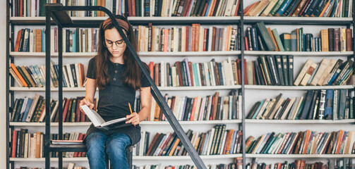 Long haired teenage girl with glasses reads textbook preparing for tests while brunette woman takes...