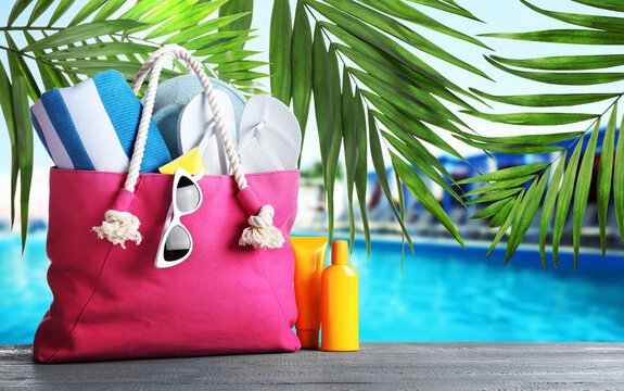 Bag with different beach accessories on grey wooden surface and green palm leaves near outdoor swimming pool