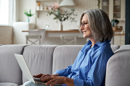 Happy 60s older mature middle aged adult woman holding laptop computer sitting on couch at home. Smiling beautiful elegant mature senior grey-haired lady using technology device in living room.