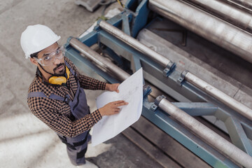 High angle view of heavy industry worker with safety headphones and hard hat in industrial factory holding blueprints