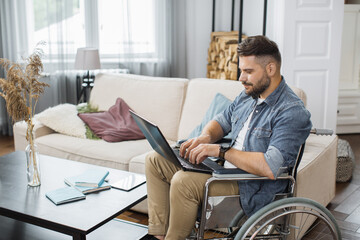 Side view of concentrated young man in wheelchair typing on wireless laptop in bright living room. Caucasian male employee with disability working at international company from home.