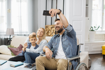 Male wheelchair user posing on smartphone camera with happy wife and school age son in modern...