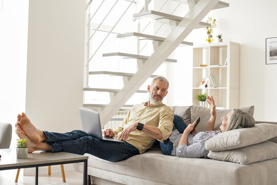 Happy mature older family couple talking using devices relaxing at home on couch. Smiling middle age senior husband and wife having conversation resting holding laptop and phone in modern living room.