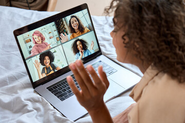 Fototapeta premium Young African American teen girl lying on bed having virtual meeting waving hand chatting with friends during online video call at home having fun using laptop computer. Over shoulder view