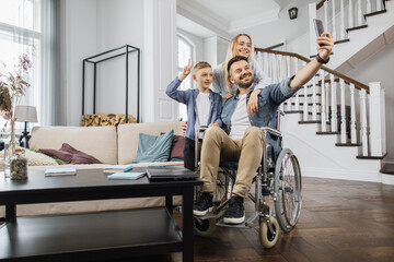 Young man sitting in wheelchair while shooting selfie on modern smartphone with wife and son standing near. Happy caucasian family smiling and looking at device camera in bright living room.