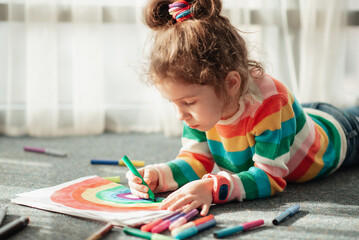 A cute little girl sits on the floor in the rays of the sun and draws a rainbow with colorful...