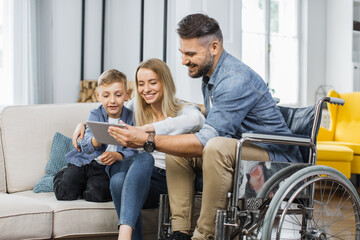 Caucasian man in wheelchair and his lovely wife with cute son watching video on digital tablet at home. Concept of family, disability and technology.