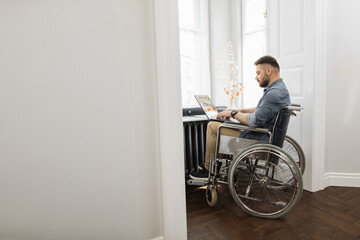 Fototapeta na wymiar Serious disabled young man using laptop for remote job at home. Confident caucasian male in wheelchair surfing internet near window at workplace.