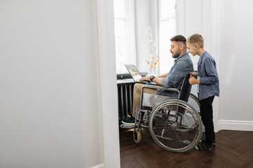 Fototapeta na wymiar Lovely son standing behind dad that sitting in wheelchair with modern laptop on knees. Caucasian bearded man and school age boy spending time together at home.