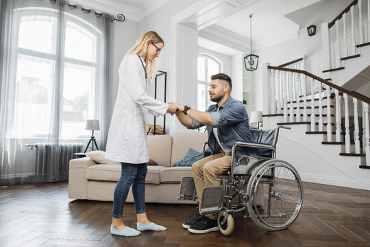 Positive young therapist assisting handsome bearded man to stand up from wheelchair at cozy home. Satisfied man beginning to feel muscles in legs while trying to walk.