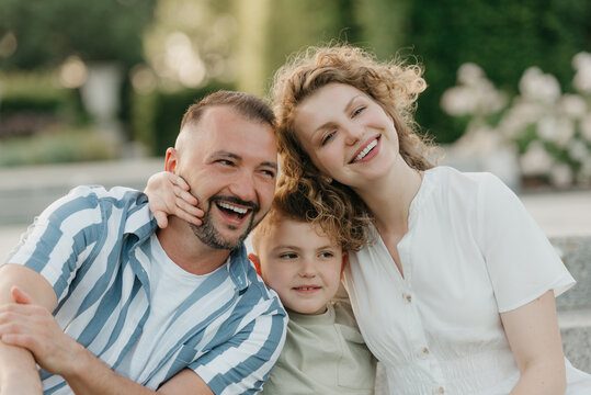 Father, mother, and son are sitting on the steps in the garden of an old European town. A close photo of a laughing family in the park in summer at sunset.
