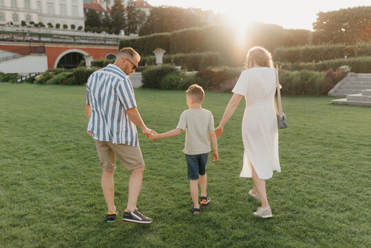 A photo from the back of a family is strolling the garden of the palace in an old European town. A happy father, mother, and son are holding hands in the evening.
