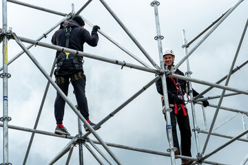 An industrial climber wearing a helmet and a protective belt is tied with a rope to a support at a high altitude. A specialist assembles a stage structure from a modular system of metal scaffolding.