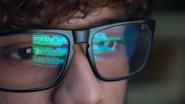 Focused indian developer coder wears glasses working on computer looking at programming code data cyber security digital tech reflecting in spectacles developing software program, close up view.