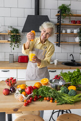 Attractive senior woman drinking freshly squeezed orange juice, looking happy and healthy. Concept of active lifestyle of mature people, cooking organic food. Mature female at home at her kitchen