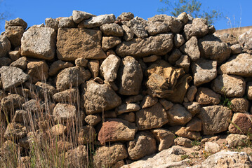 An ancient wall of rough stones stacked on top of each other in the traditional way in Sicily. Above is blue sky. There is dry grass in front of the wall.