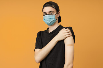 A young man in two medical masks after vaccination shows a medical plaster on his arm, a portrait on a yellow background. The concept of vaccination against coronavirus.