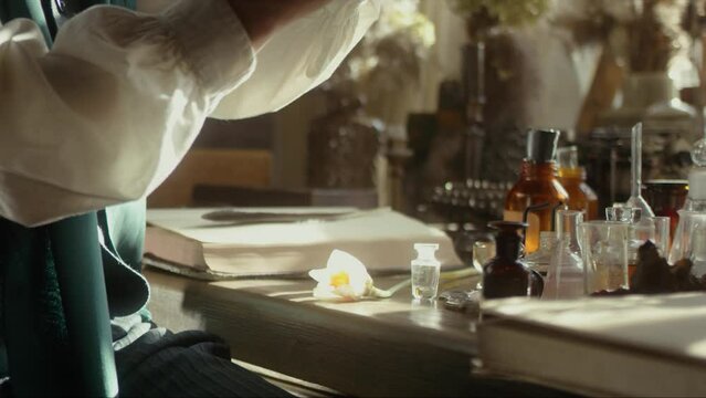 A close-up of a perfumer at his desk looking for a new fragrance. Vintage cinematic perfumery concept. Sunny day in the working workshop. Lots of ingredients, glass flasks. Depth of field. 4K video