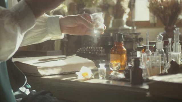 A close-up of a perfumer at his desk looking for a new fragrance. Vintage cinematic perfumery concept. Sunny day in the working workshop. Lots of ingredients, glass flasks. Depth of field. 4K video