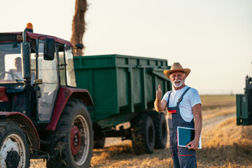 Obraz premium Senior agronomist and farm owner is standing in front of a combine unloading wheat into a tractor trailer. He is smiling and showing thumbs up.