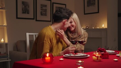 Happy smiling young couple in love hugging and laughing, drinking wine, talking, celebrating Valentines day dining together, having romantic dinner date in candle light sitting at home table. 