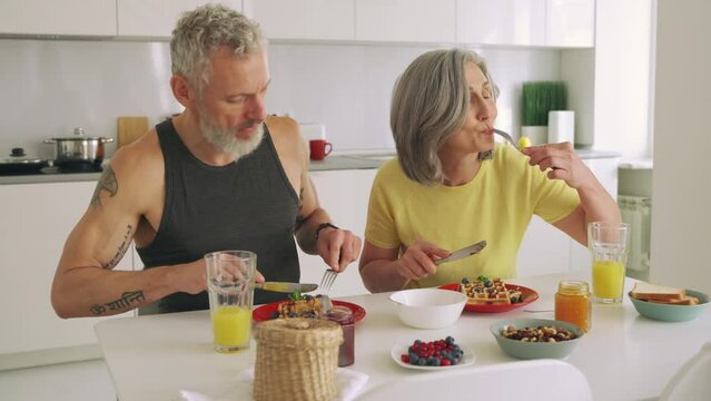 Happy healthy fit mature older family couple having breakfast sitting at kitchen table. Smiling mid age 50s husband and wife talking while eating waffles, having fun enjoying morning meal together. 