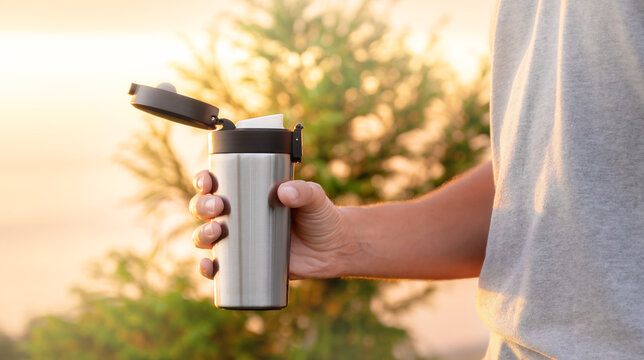 Man traveller holds silver thermos cup, sunset sky on the background, concept of outdoor vacation, space for text