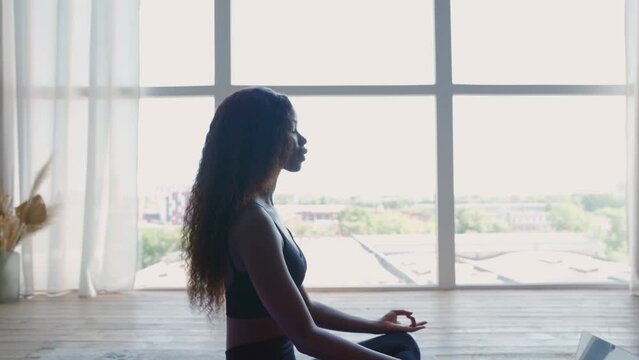 Young calm fit African black woman sitting on floor at home near window doing yoga breathing exercise, meditating learning online training virtual class with teacher on computer. Mental health concept