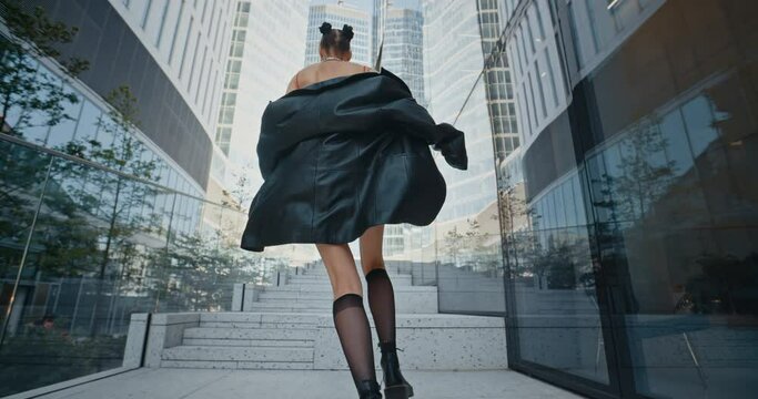 Stylish girl wearing black leather jacket and short skirt runs up stairs next to high rise glass buildings in downtown business center in modern city. Fashionable female, summer day, slow motion