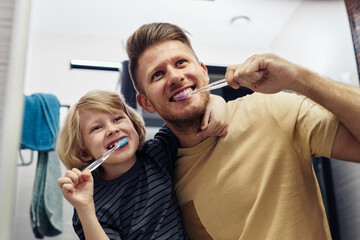 Low angle portrait of playful father and son brushing teeth together in morning and looking in...