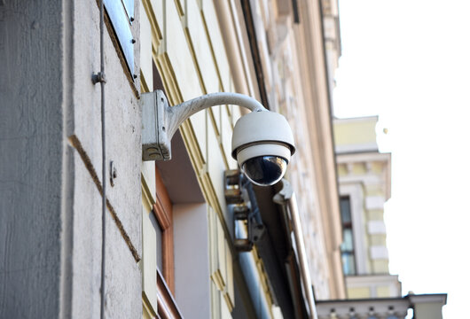 Mounted outdoor surveillance camera of round shape attached to the wall of the building from the street side on a clear day