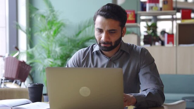Young indian concentrated ceo sitting at desk using laptop in office. Serious financial analyst working with big data. Business technologies concept