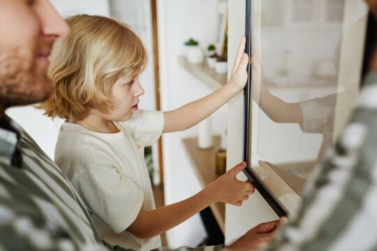 Side view portrait of cute blonde boy helping father hanging picture on wall while decorating new house