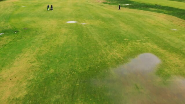 Drone shot of a wide flat green golf course with big puddles. Some people moving their golf equipment to the next hole. . High quality 4k footage