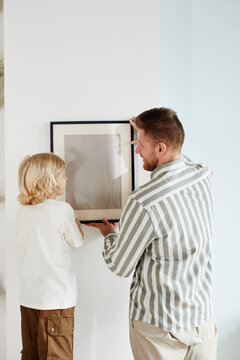 Vertical back view portrait of father and son hanging picture together and looking at each other