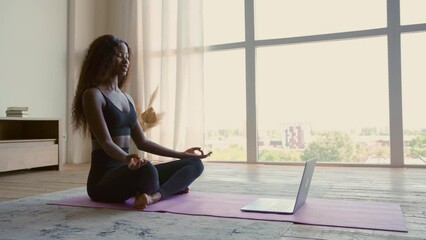 Young calm fit healthy African black woman sitting on floor at home doing yoga breathing exercise, meditating learning online training virtual class on computer. Exercises for mental health concept. - Powered by Adobe