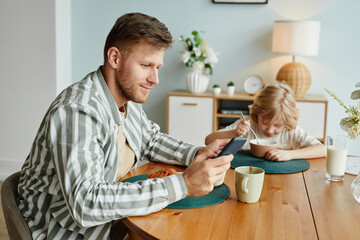 Fototapeta premium Side view portrait of father using smartphone at dining table while enjoying breakfast with son