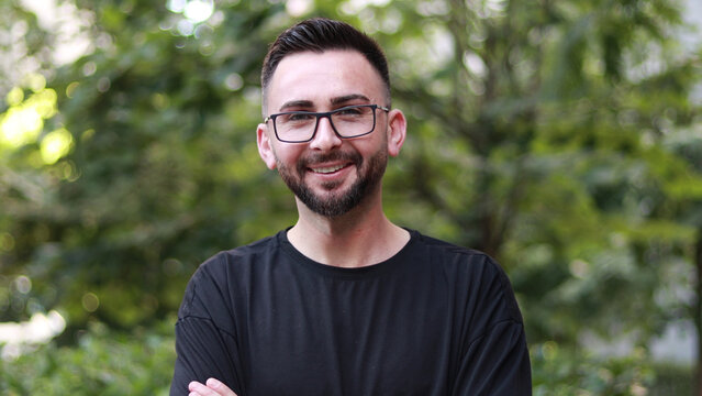 Portrait of young handsome man with beard and glasses. Young man in black shirt smiling looking at camera. Outdoor nature background